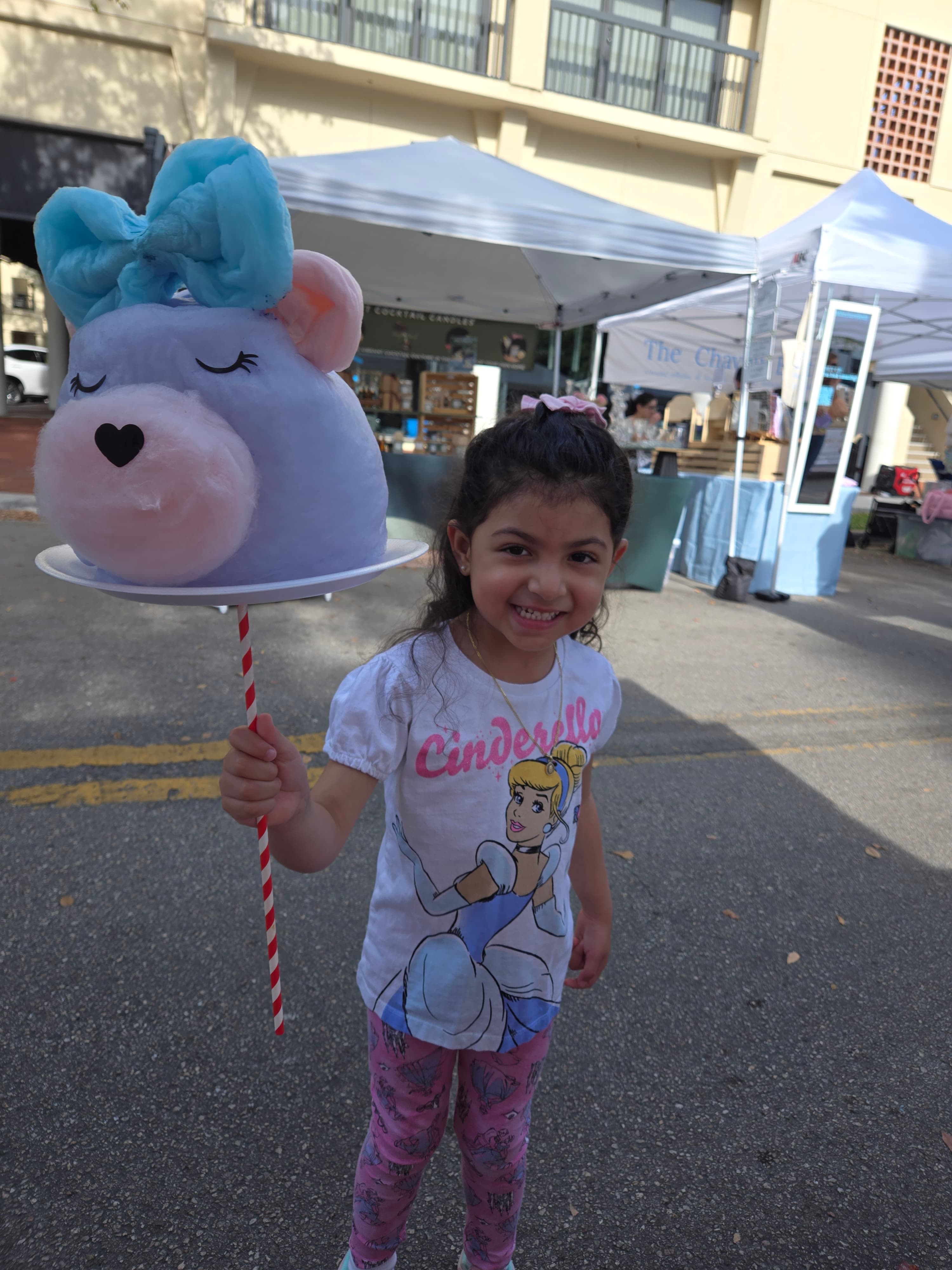 Smiling young girl holds a large, blue character-shaped cotton candy at an outdoor fair.