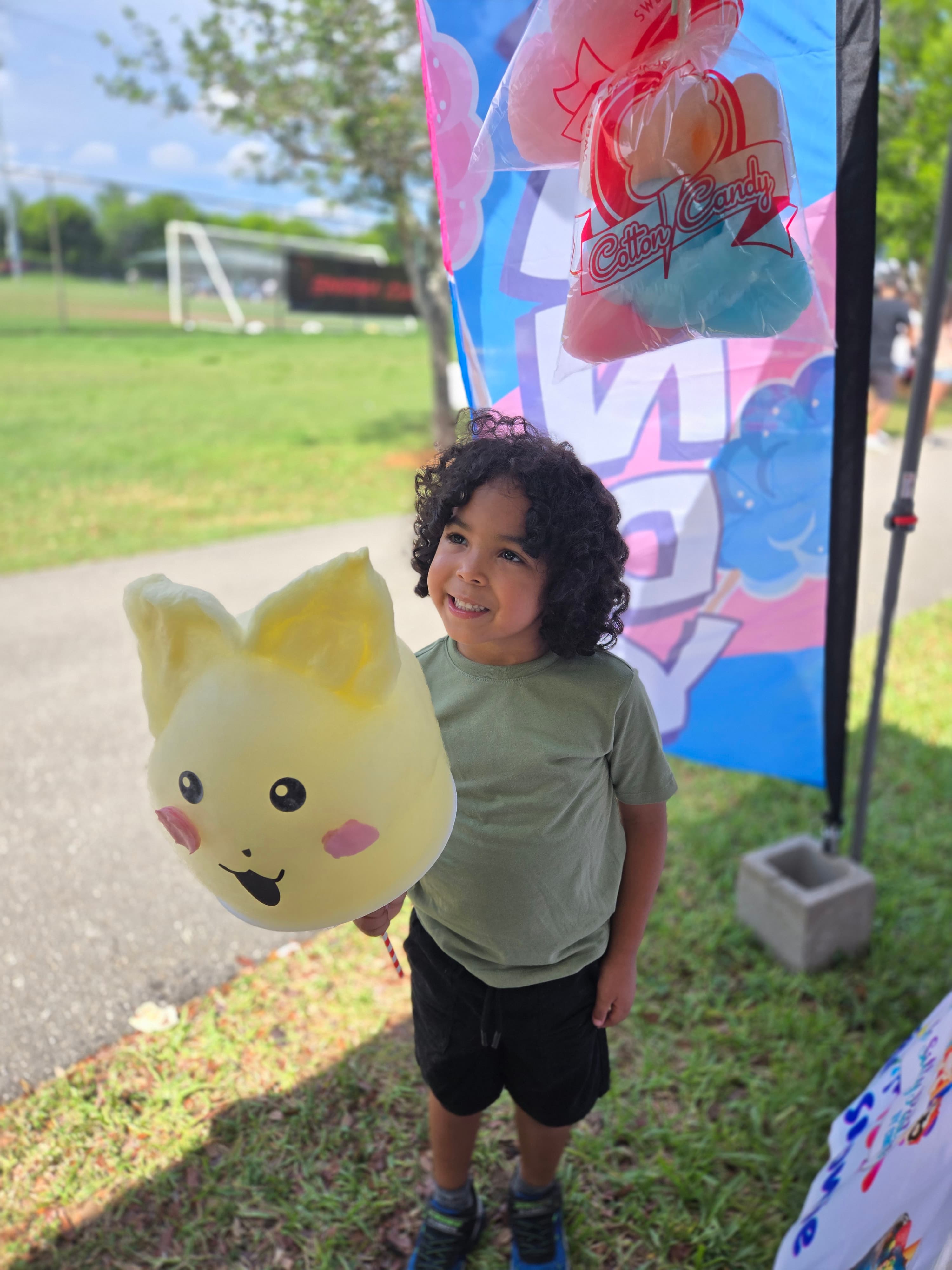 Smiling child with curly hair holding a large yellow character-shaped cotton candy outdoors.