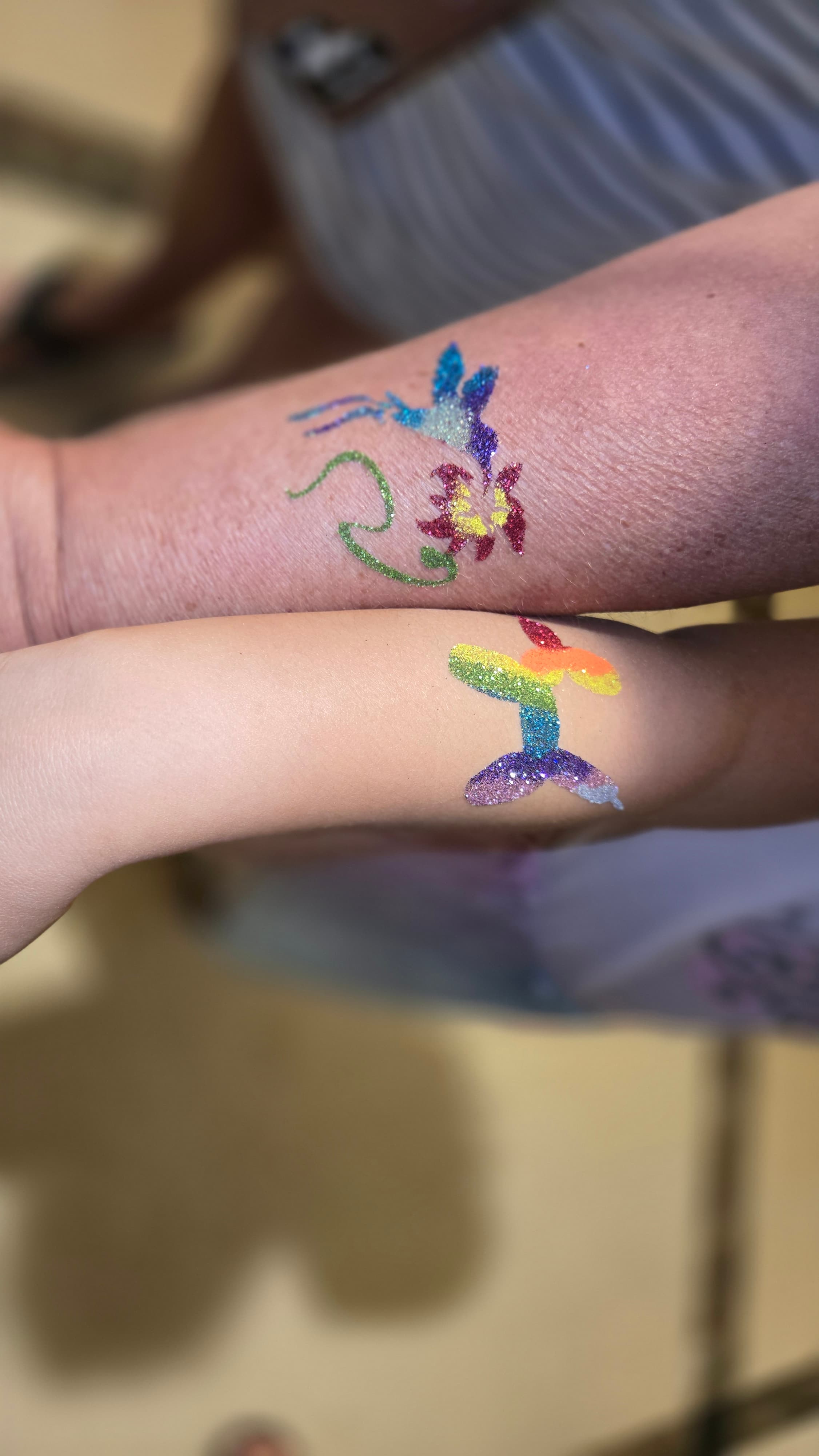 Two arms featuring vibrant glitter tattoos of a hummingbird, flower, and rainbow balloon dog.